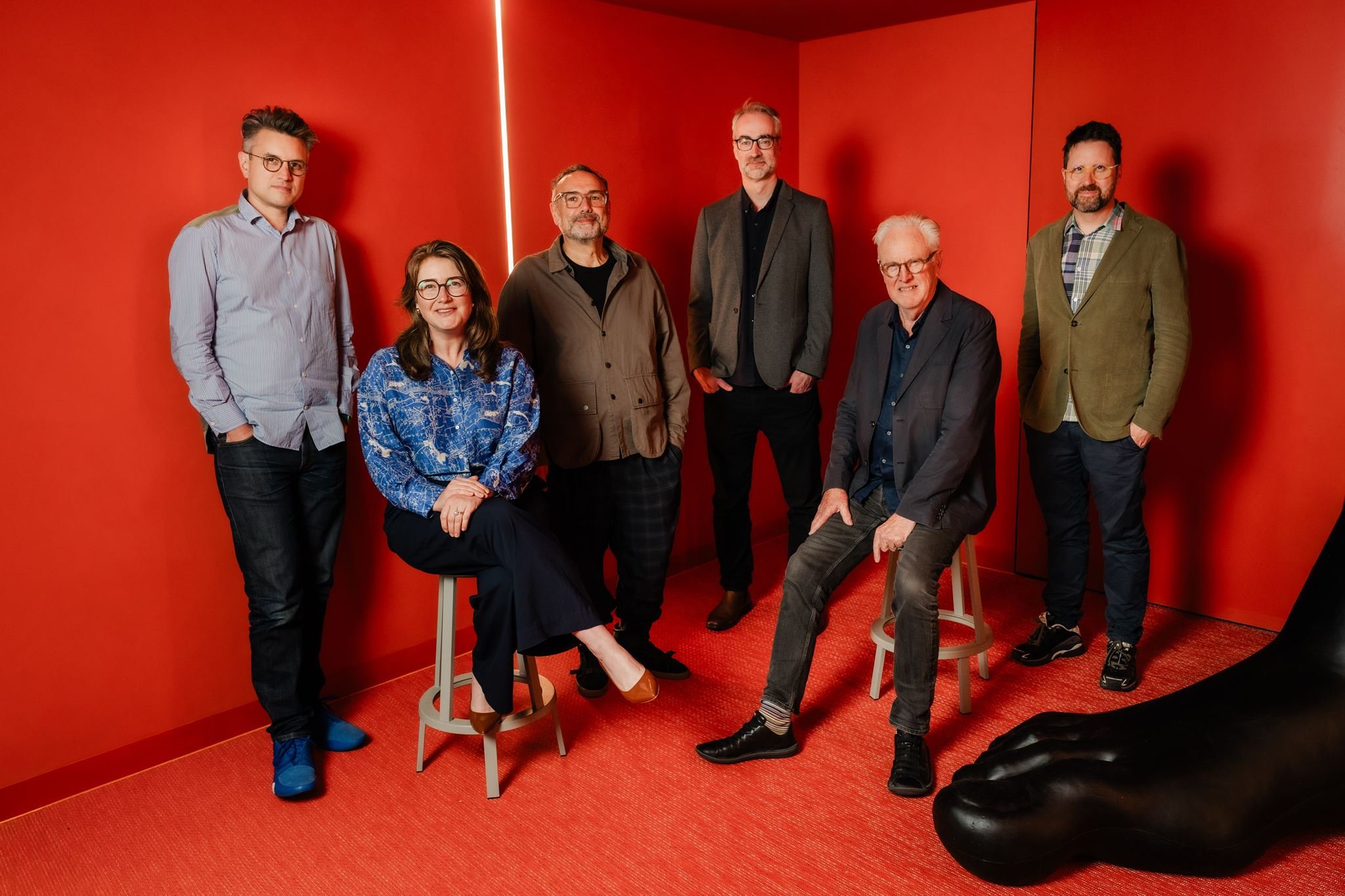 Seven people, four men and three women, posing in a red room with minimalist decor. Some are standing, others seated on stools; they are dressed in casual to business casual attire.