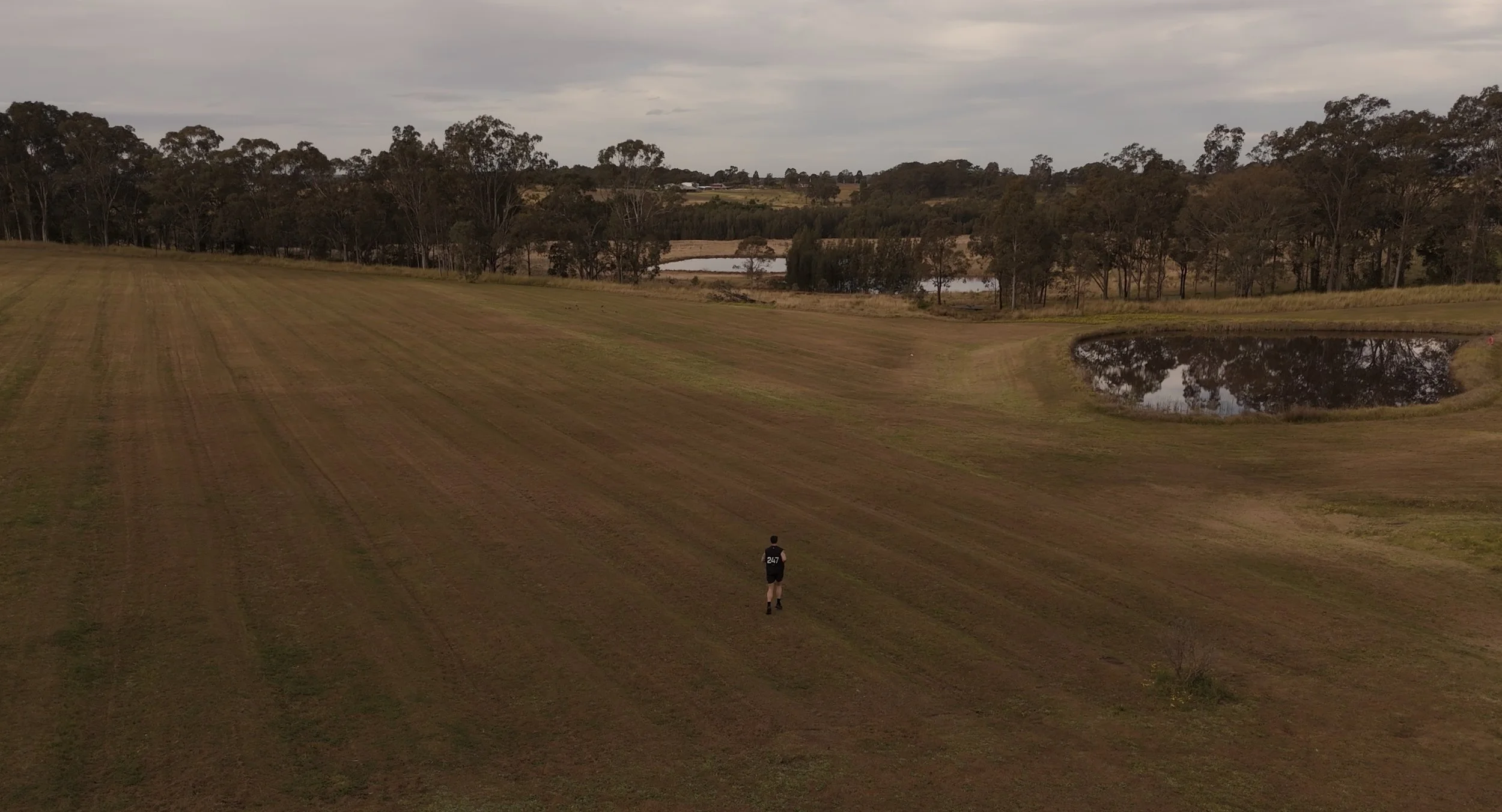 A person running on a grassy field with trees, ponds, and a cloudy sky in the background.