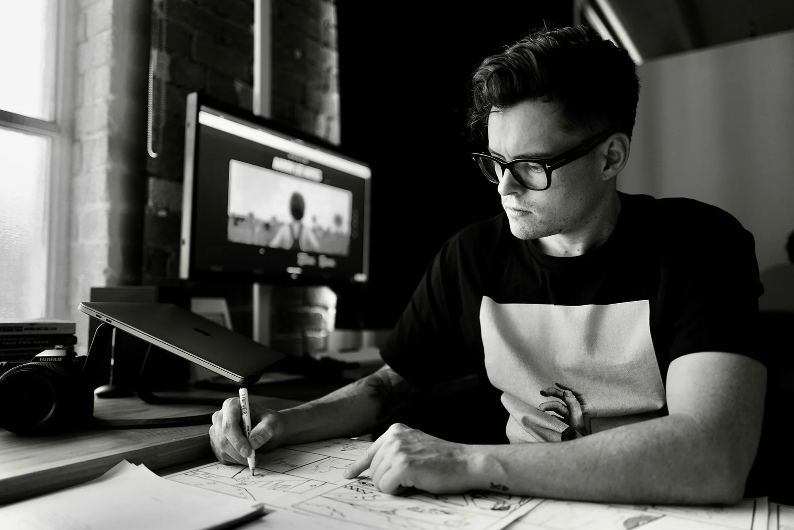 A man with glasses drawing on papers spread out on a desk, with a computer monitor and camera nearby, in a dimly lit room.