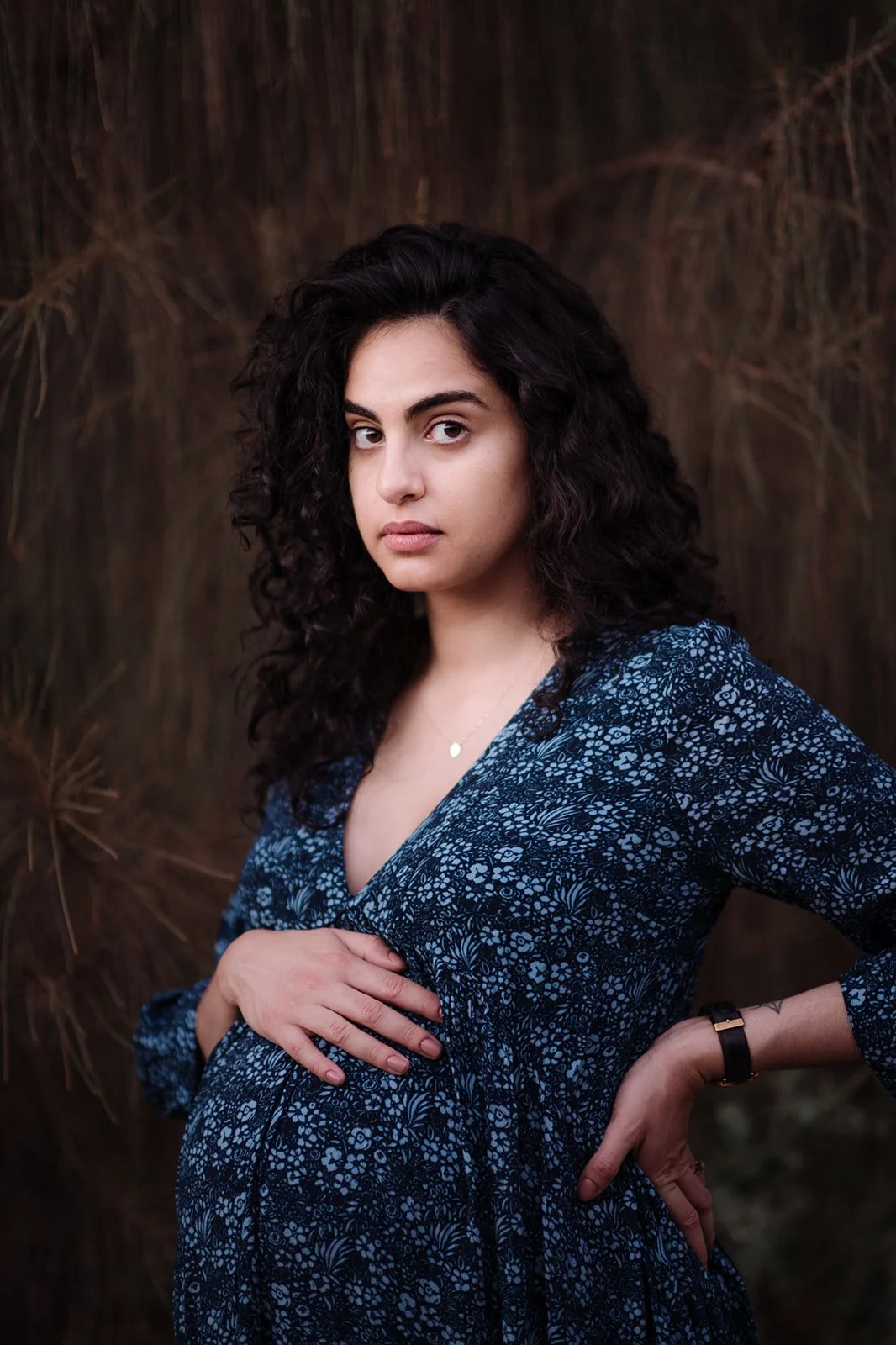 A woman with dark curly hair wearing a blue patterned dress standing outdoors against a background of dry grass or plants, looking at the camera with a serious expression.