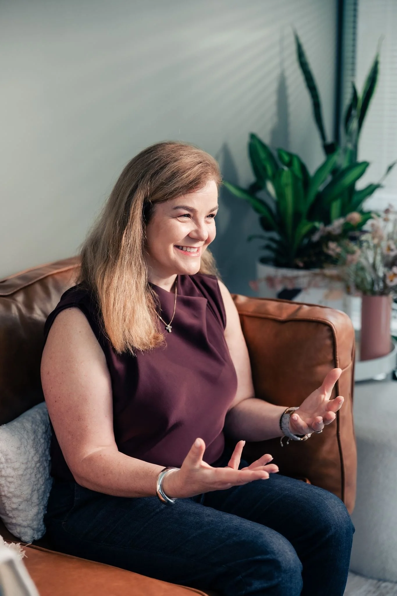 A woman with long blonde hair smiling and gesturing with her hands while sitting on a brown leather couch in a room with a large green plant and window in the background.