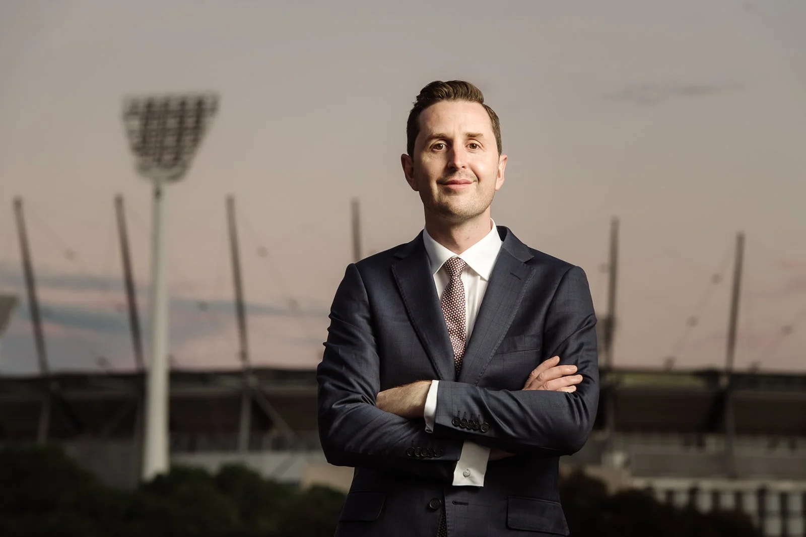 A man in a suit standing with arms crossed in front of a stadium with bright stadium lights.