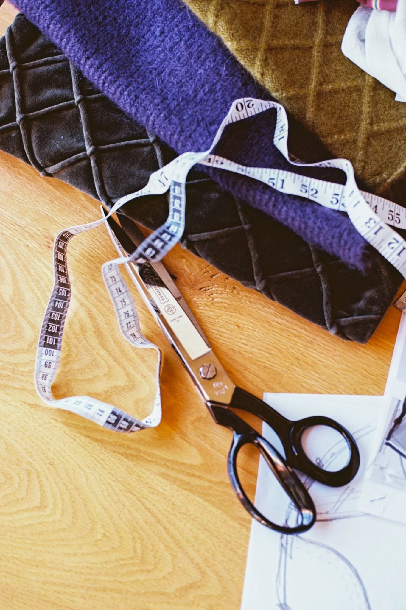 A pair of black scissors, a measuring tape, a piece of paper with a drawing, and some fabrics on a wooden table.