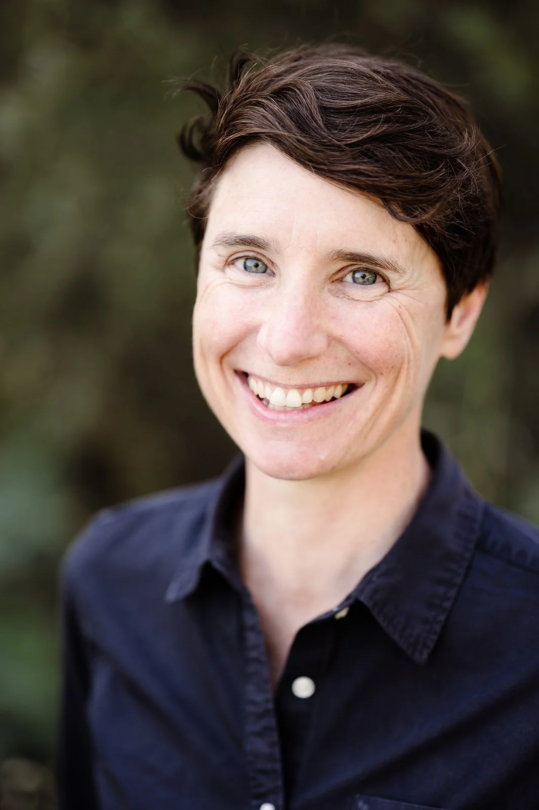 Close-up portrait of a smiling woman with short dark hair, blue eyes, and wearing a dark collared shirt, outdoors with blurred green background.