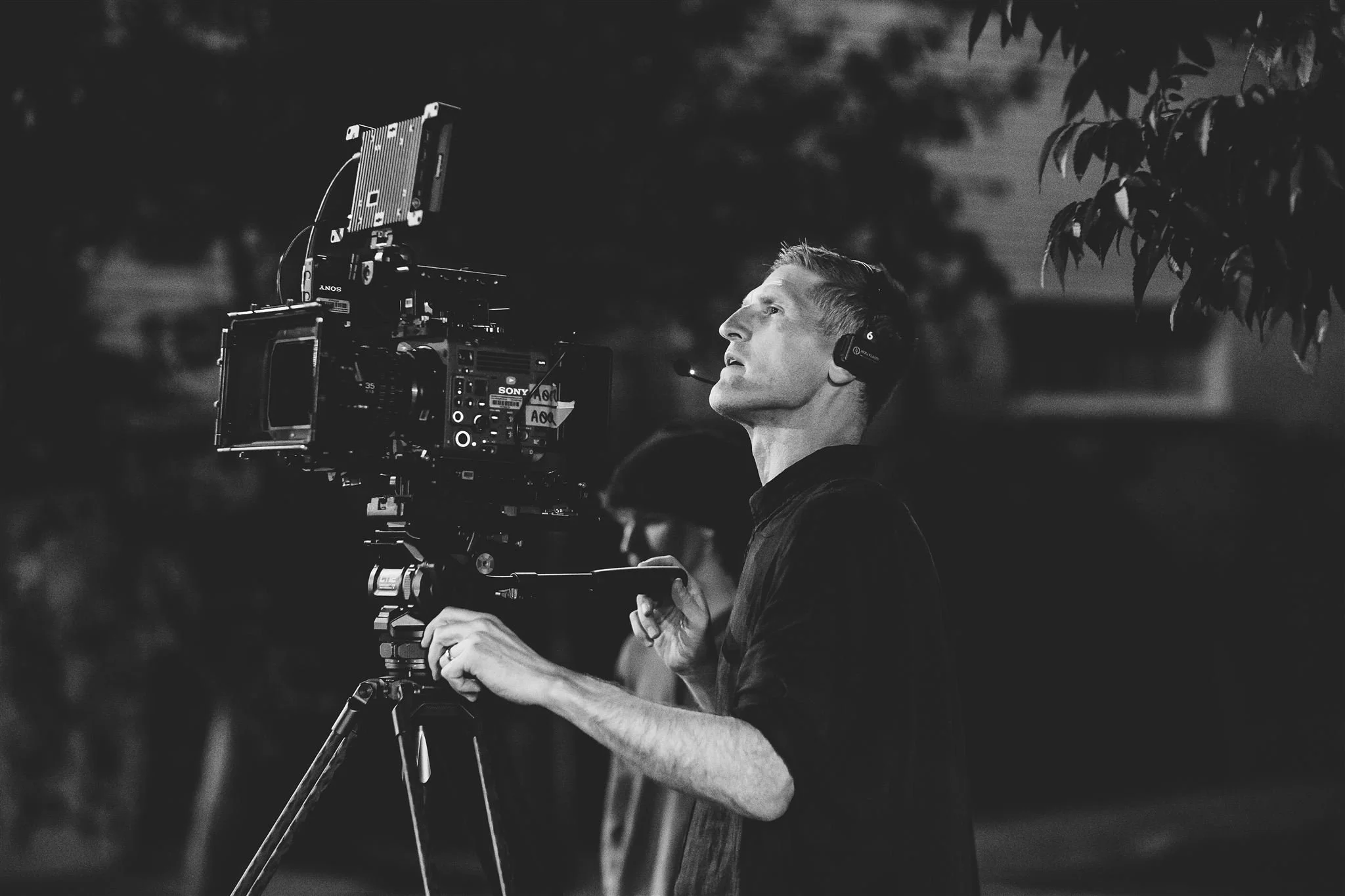 A man operating a professional camera on a tripod, wearing headphones, in an outdoor setting at night.