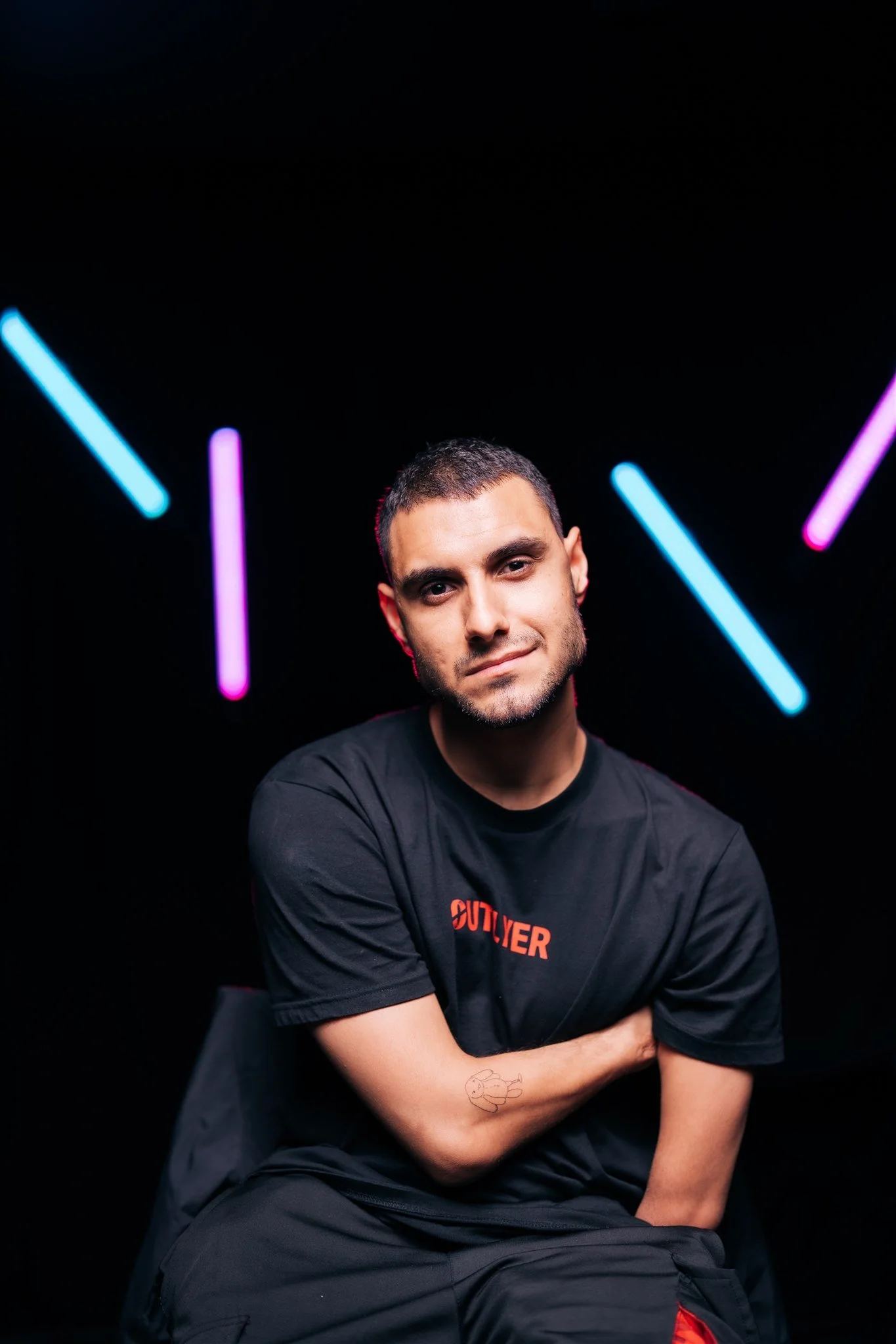 Portrait of Emmanuel Kelly, seated with arms crossed against a black background with neon-coloured light bars.