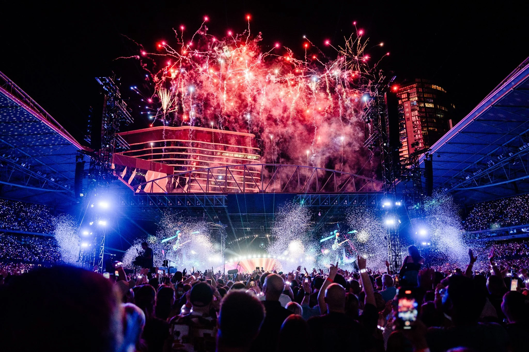 Crowd watching fireworks light up the night sky during a concert or festival at a stadium or outdoor venue.
