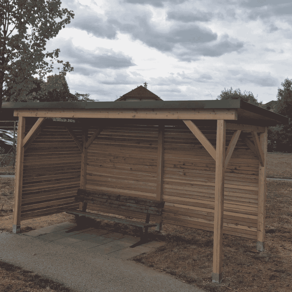 Eine neue, halboffene Bushaltestelle mit Holzlatten und einer Bank davor, im Freien mit Bäumen und einem Haus im Hintergrund bei bewölktem Himmel.