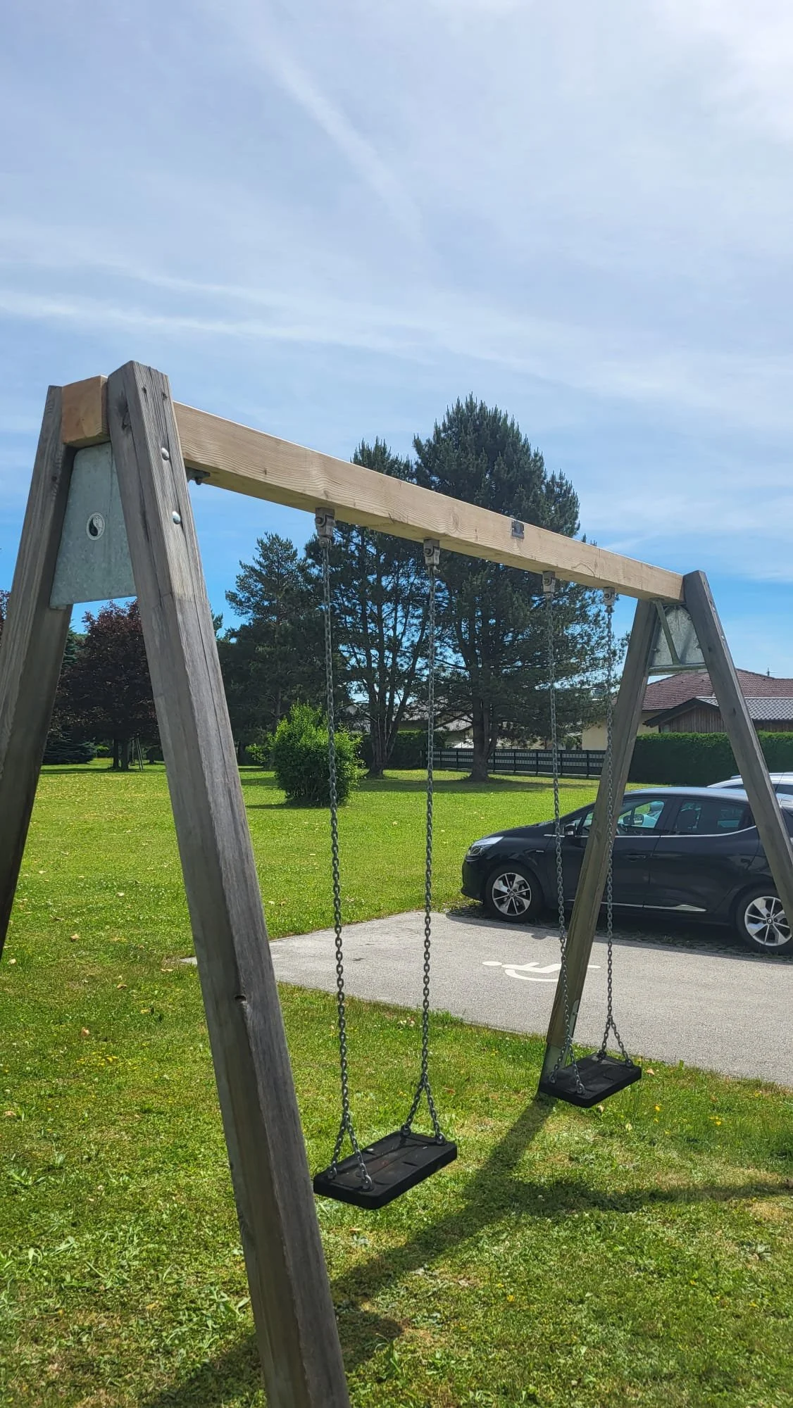 Schaukelsitz auf einem Kinderspielplatz, ein Auto parkt im Hintergrund, Bäume und ein Haus sind zu sehen.