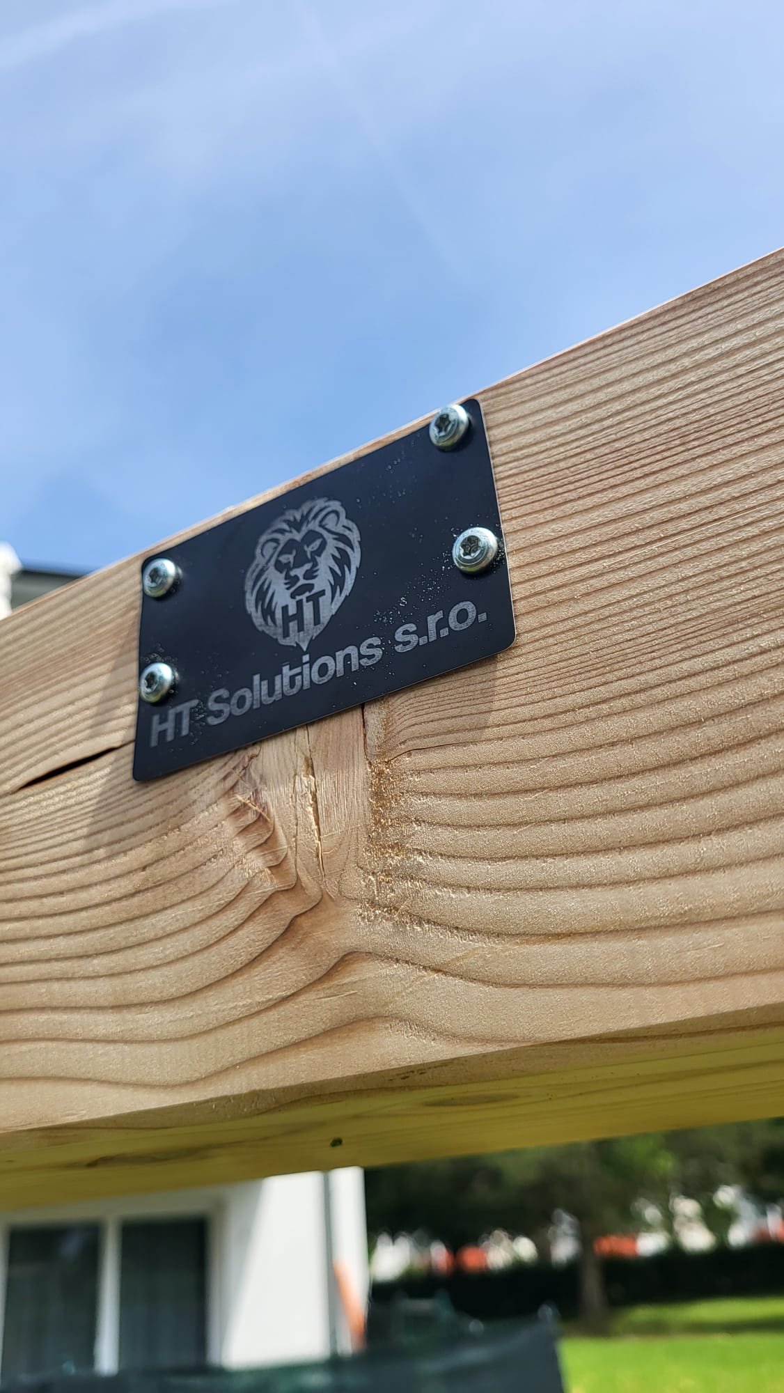 Nahaufnahme eines Holzbretts mit einem schwarzen Metallschild, auf dem das Logo eines Löwen und der Text 'HT Solutions s.r.o.' zu sehen sind.
