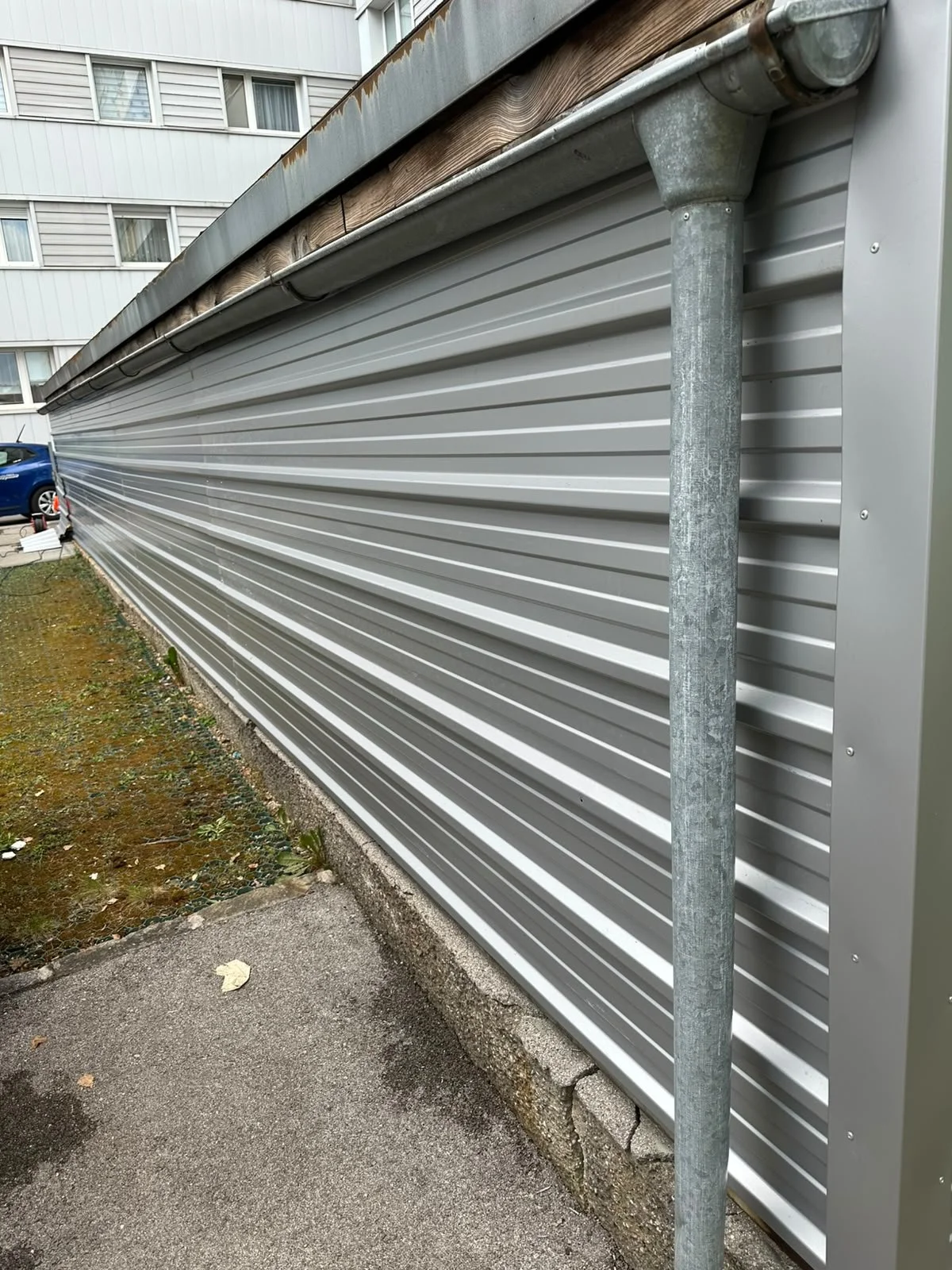 Außenansicht eines Gebäudes mit einer metallischen Rollladentür, einem Regenfallrohr und einer kleinen befestigten Bodenfläche, umliegendes Grünzeug und Fahrzeuge im Hintergrund.