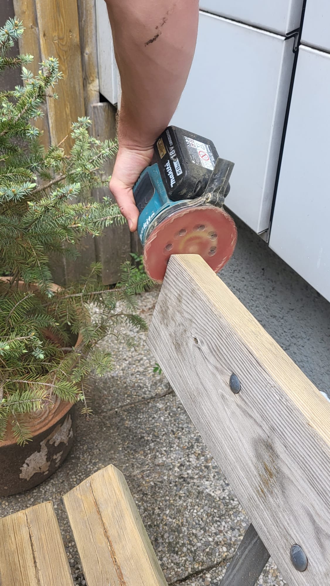 Person verwendet eine elektrische Schleifmaschine, um eine Holzlatte zu glätten, draußen neben Pflanzen und Gartenmöbeln.