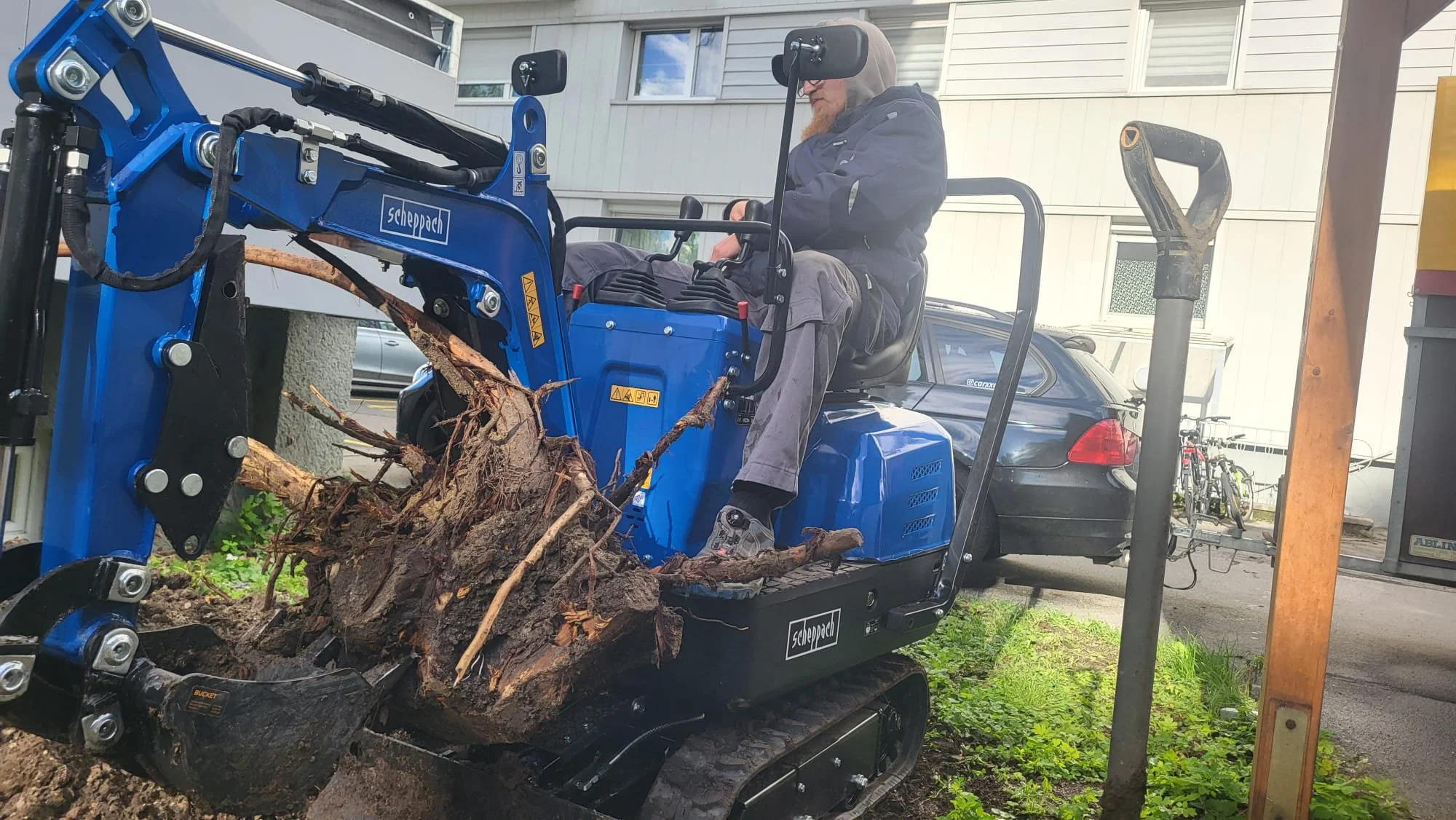 Mann steuert kleinen blauen Traktor mit Holzstämmen und Ästen auf dem Anhänger, im Hintergrund Wohnhaus, Fahrradständer mit Fahrrädern.