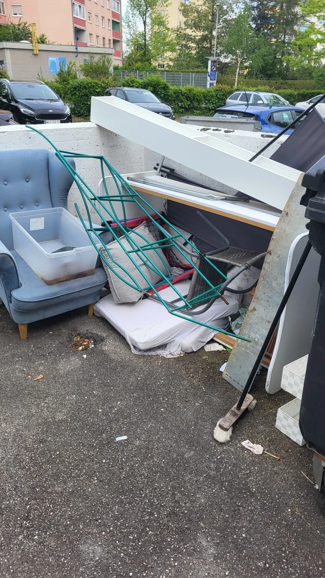 Ein Haufen von alten Möbeln und Haushaltsgegenständen im Freien, darunter ein rotes Metallbettgestell, ein graues Sofa, eine Kunststoffbox, eine weiße Anrichte und andere Gegenstände auf dem Boden und auf einer Mauer.