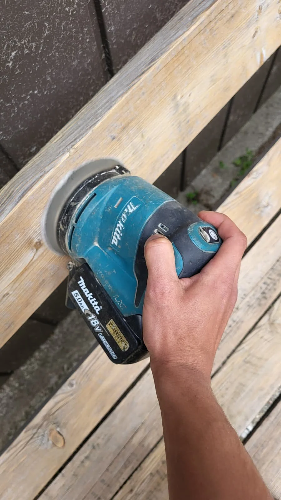 Eine Hand hält ein blauer Makita-Schleifer, der auf Holzoberfläche arbeitet.