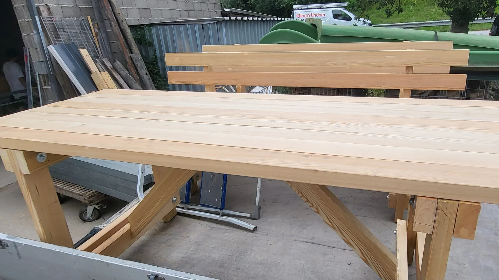 Holztisch mit Holzbank, im Hintergrund Baustoffe, Paletten, Fahrräder und ein Transporter.