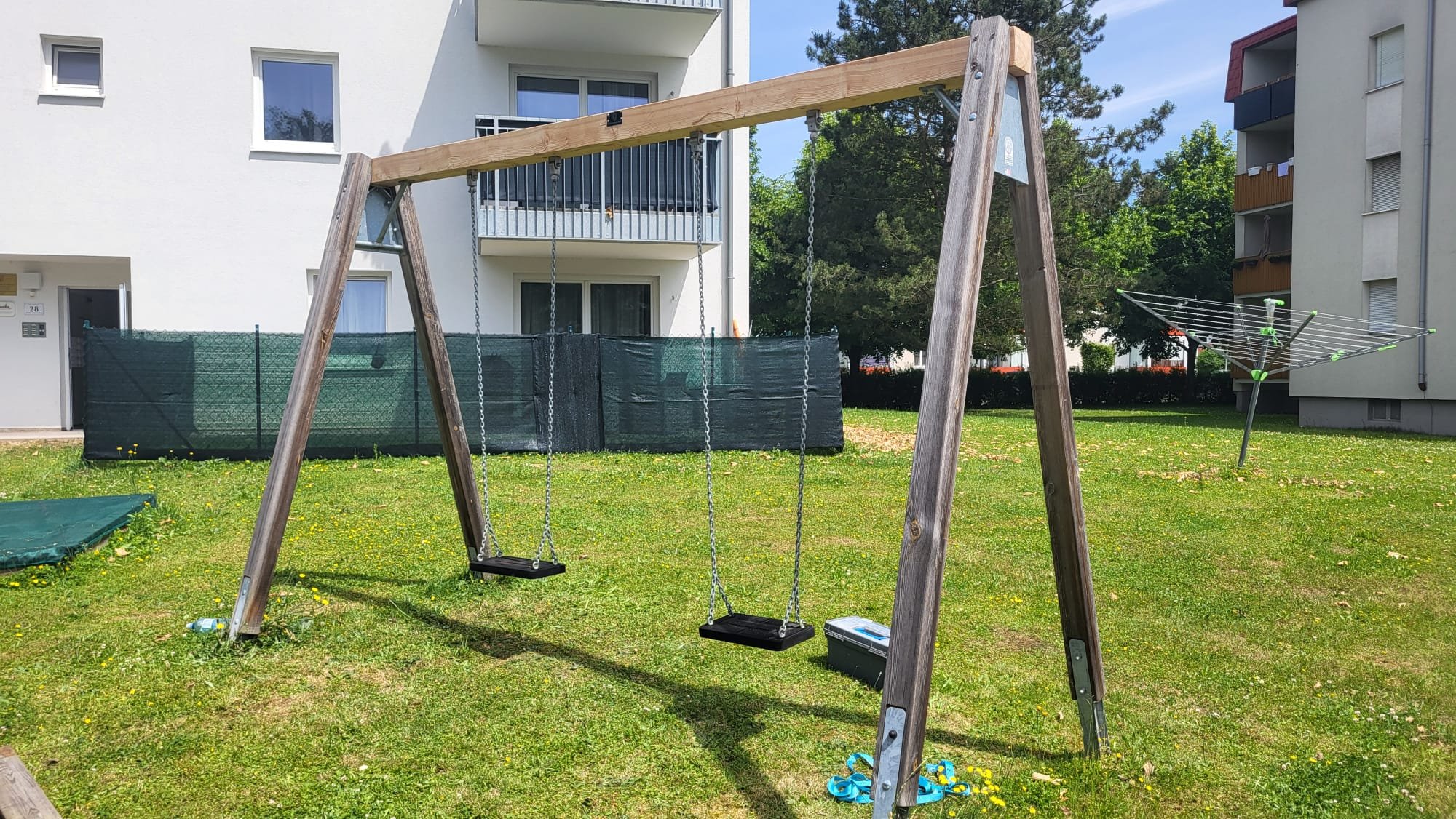 Kinderspielplatz mit einer Schaukel, einem Wäscheleine-Trockner und einem grünen Rasen in einer Wohnanlage.