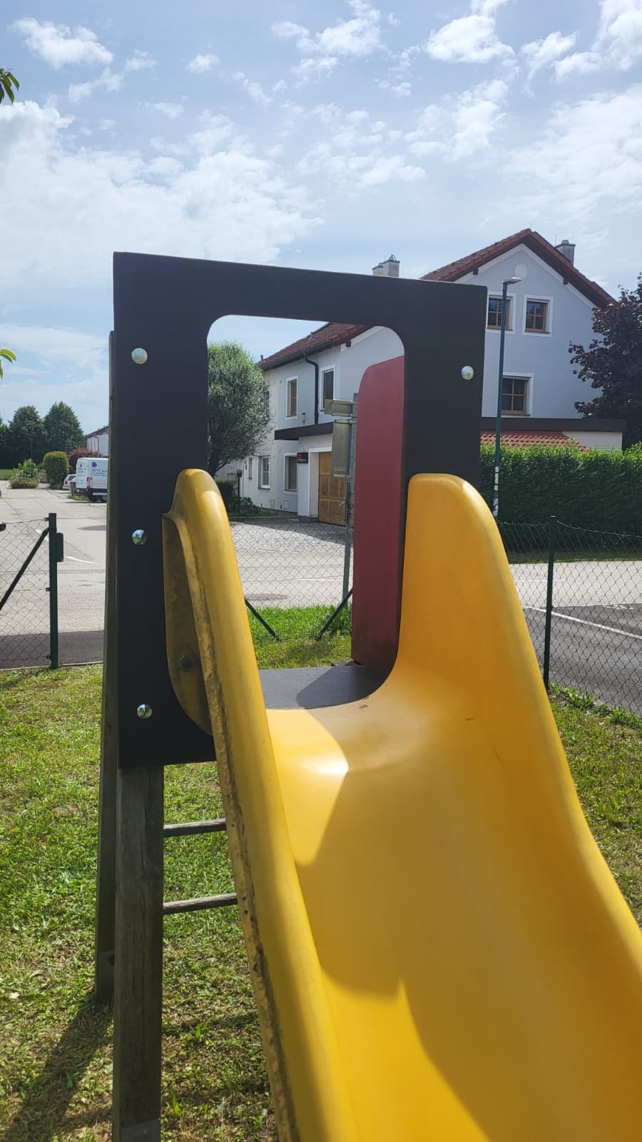 Blick durch eine Kinderrutsche mit einem schwarzen Rahmen, gelbem Rutschenteil, rotem Sichtschutz, im Außenbereich vor Wohnhäusern, grüner Wiese und Himmel mit Wolken.