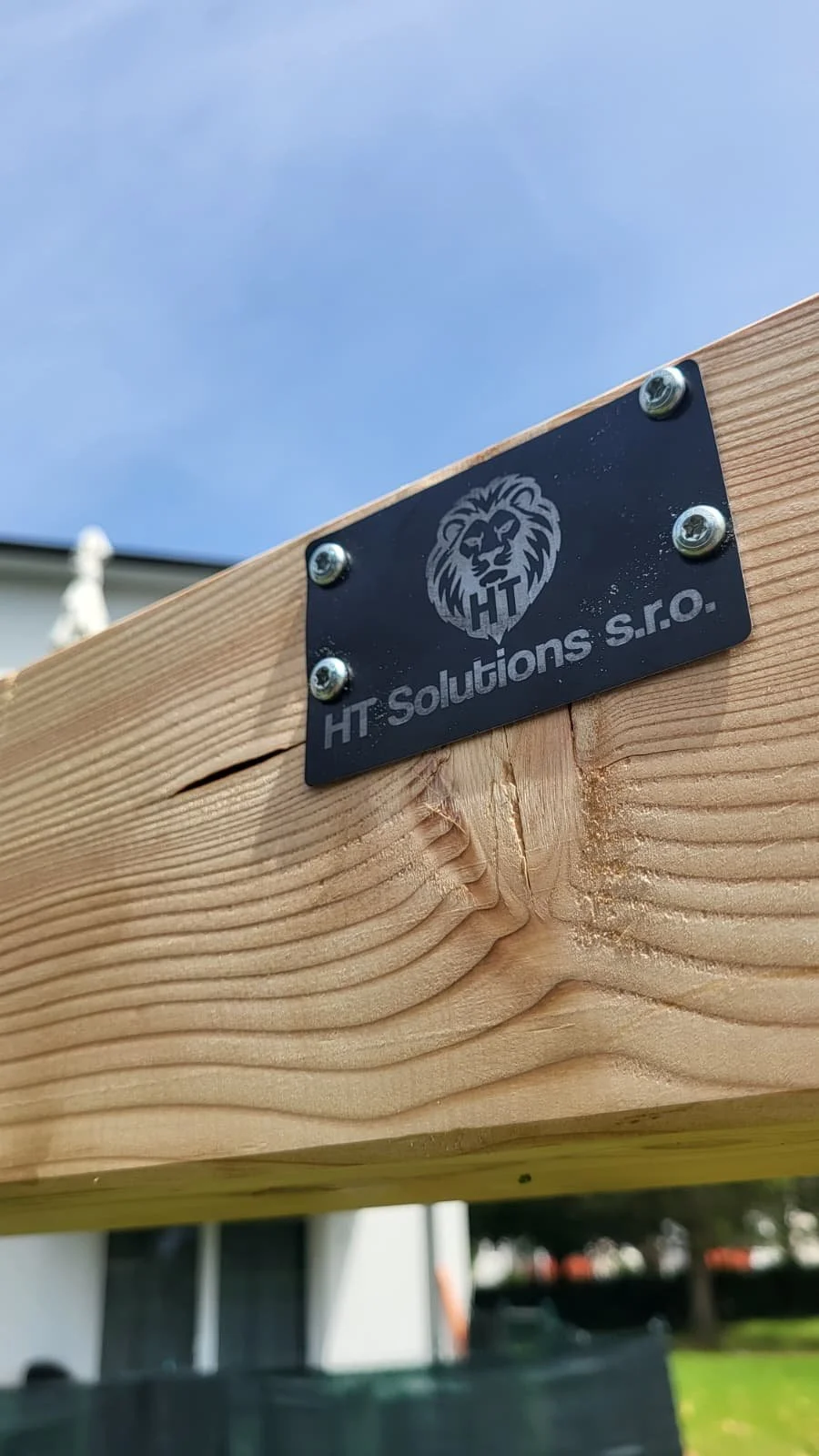 Metallplakette mit Löwenkopf-Logo und der Aufschrift "HT Solutions s.r.o." an einem Holzbalken, im Hintergrund blauer Himmel.