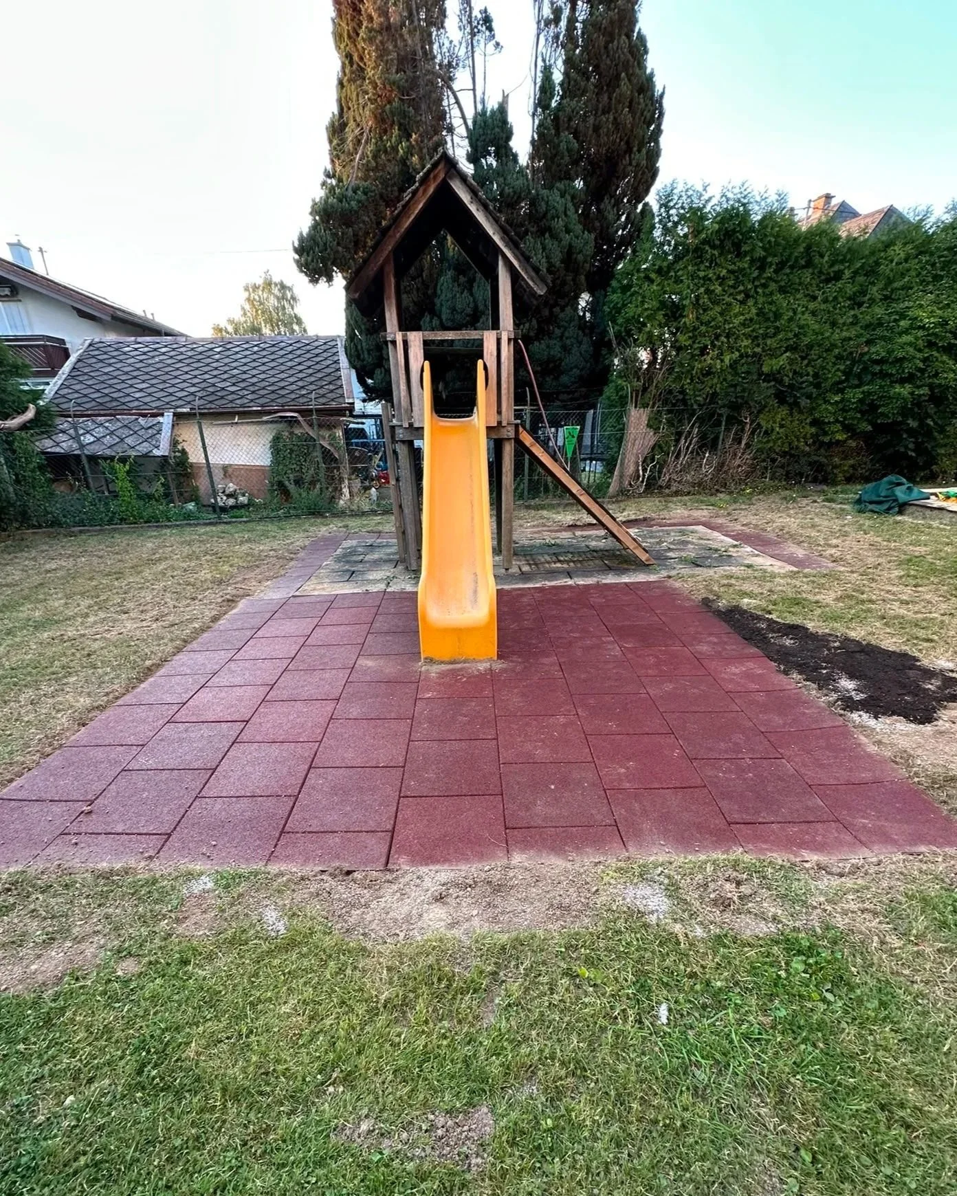 Ein Holzspielturm mit Rutsche auf einer Mischung aus roten Bodenplatten und Gras in einem Garten, umgeben von Bäumen und Zäunen.