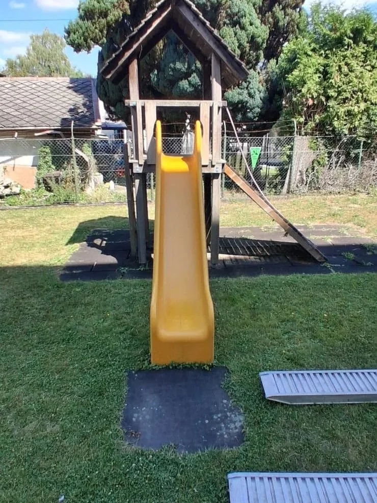 Ein kleiner Holzspielturm mit Rutsche in einem Garten, umgeben von Bäumen und Zäunen.
