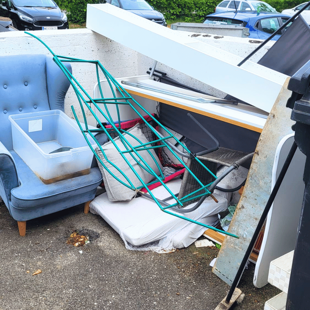 Mischmasch aus alten Möbeln und Haushaltsgegenständen im Freien, darunter ein umgestürzter Wäschekorb, eine Couch, ein Bettgestell, und andere Gegenstände.