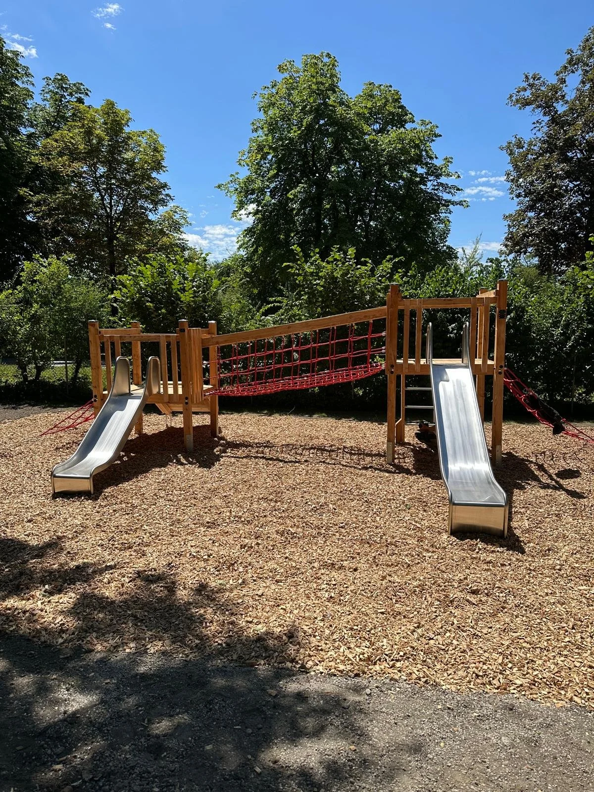 Spielplatz mit zwei Rutschen, Kletterseil und Holzstruktur, umgeben von Bäumen und einem blauen Himmel.