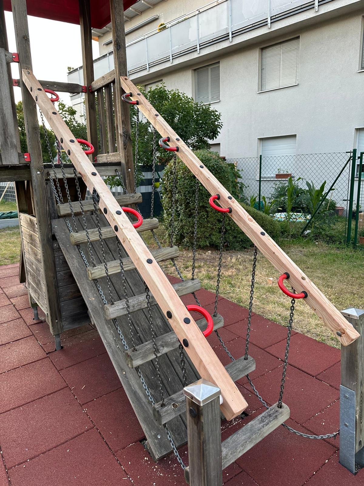 Spielgerüst für Kinder mit Kletterstangen, Sprossen und Seilen auf einer roten Gummimatte, im Hintergund eine Hauswand mit Fenstern und Fensterläden.