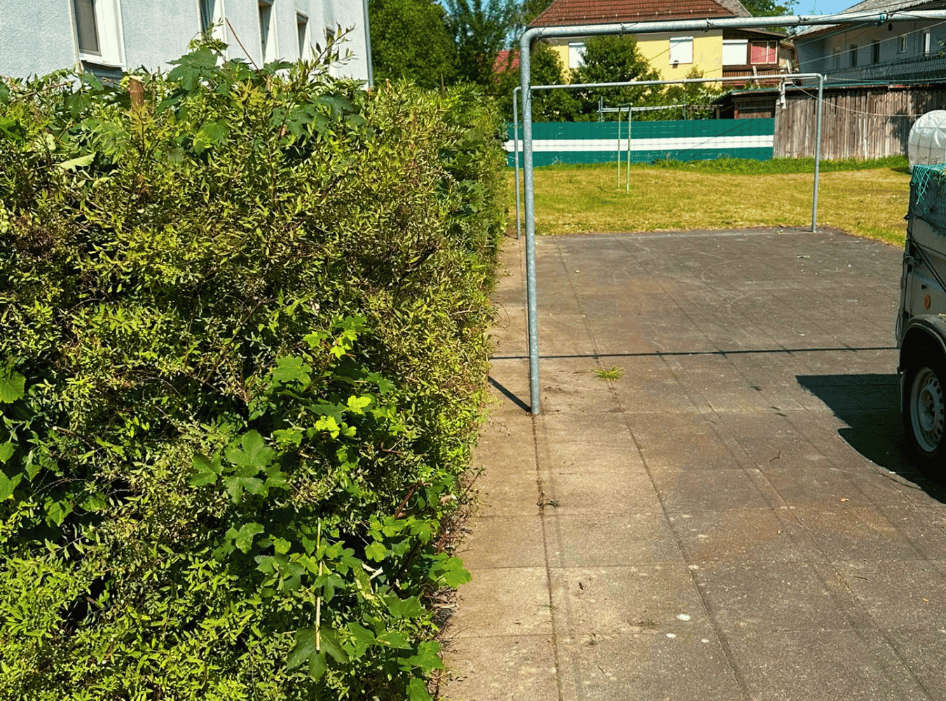 Ein gepflegter Vorgarten mit einer Hecke auf der linken Seite, einem gepflasterten Weg und einer freigehaltenen Fläche im Hintergrund mit einem Wäschelineal. Mehrere Häuser und Bäume sind im Hintergrund sichtbar.