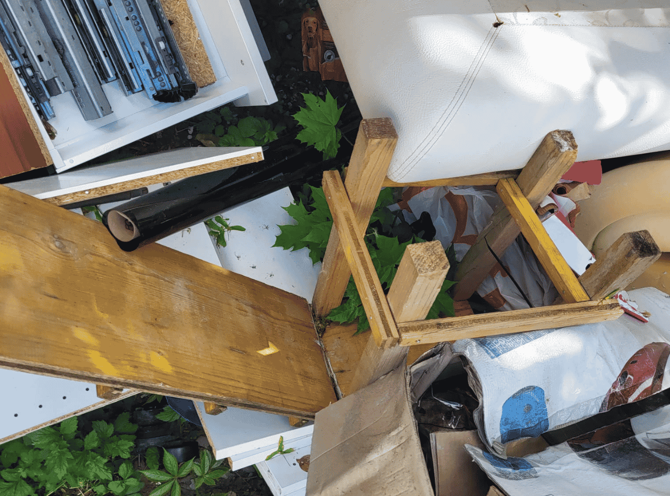 Ungeordneter Arbeitsbereich mit Holzstücken, Pflanzen, einem gemusterten Tuch, einer weißen Polsterung, einem schwarzen Rohr, einem schwarzen und weißen Regal und verschiedenen Verpackungen.