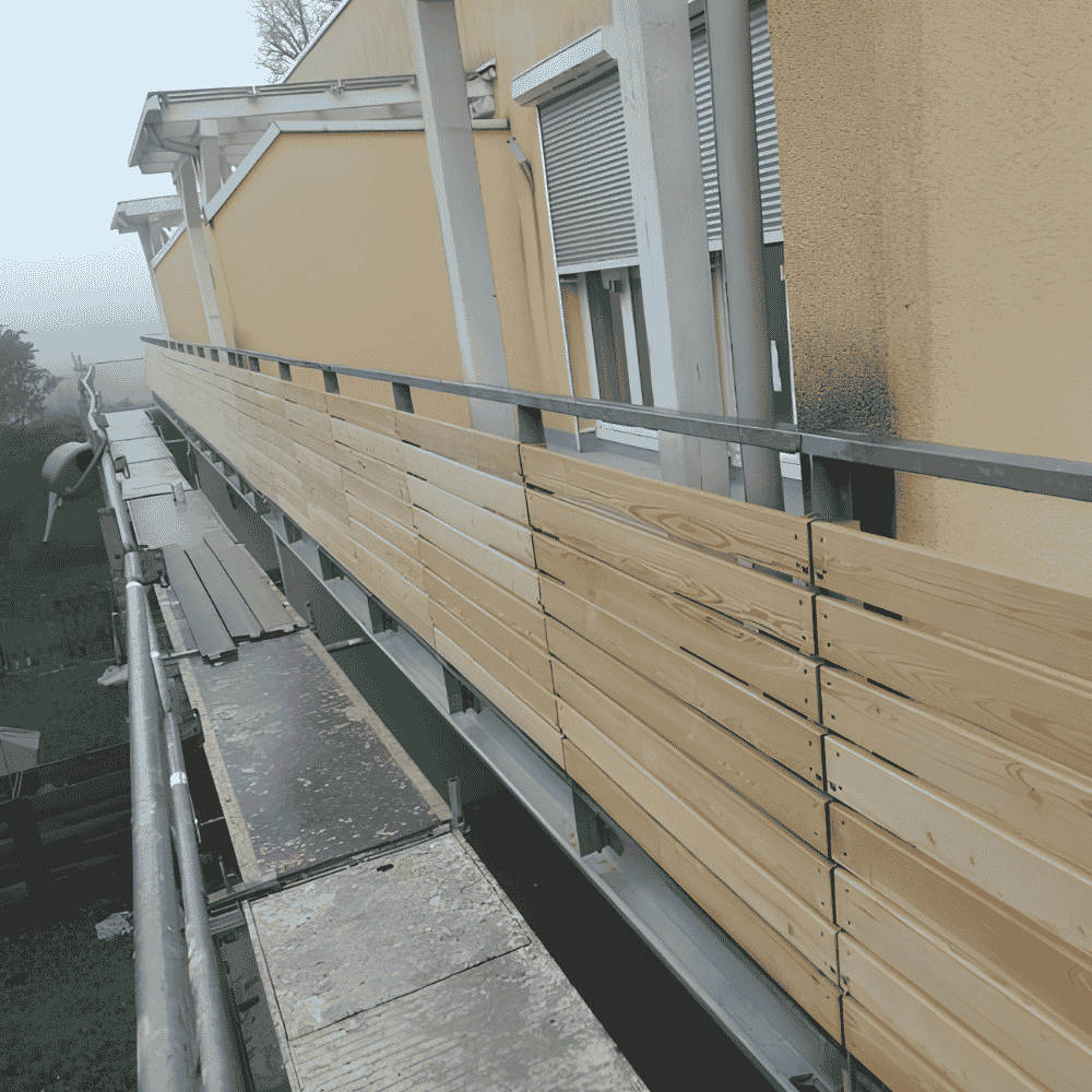 An einem Balkon wird Holzzaun installiert, das Gerüst ist sichtbar, mit einer gelben Hausfassade im Hintergrund.