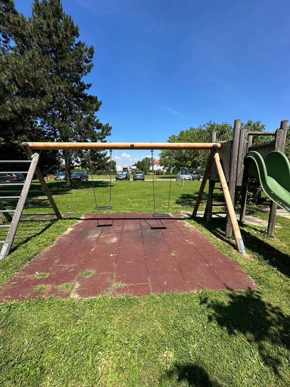 Spielplatz mit Vogelschaukeln, Rutschbahn, Kletterstange, Rasen, Bäumen und parkenden Autos im Hintergrund bei sonnigem Himmel.