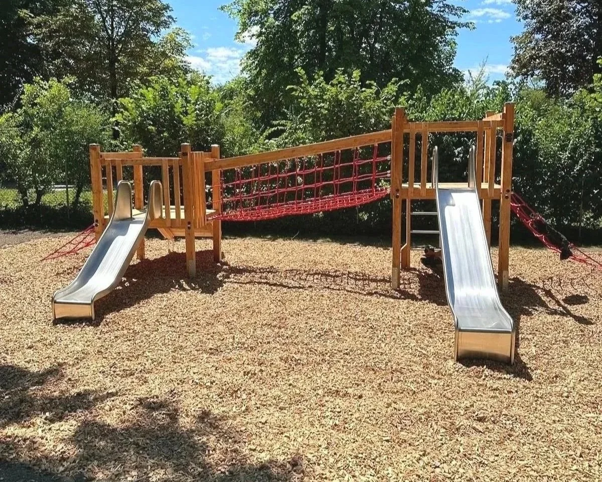 Spielplatz mit Holzrahmen, zwei Metallrutschen, rotem Netz zum Klettern, auf Sandboden, umgeben von Bäumen und Buschwerk bei Sonnenschein.
