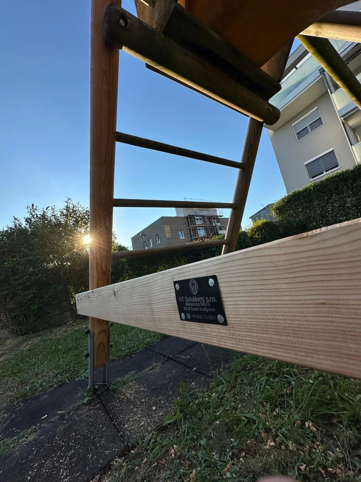 Holzleiter im Garten mit Firmenetikett, im Hintergrund moderne Wohngebäude, Sonnenuntergang bei klarem Himmel.