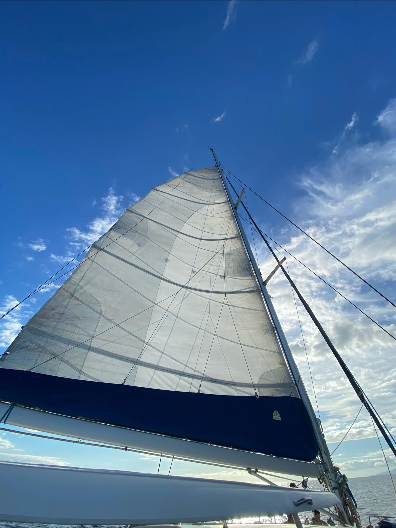 View of a sailboat with white sails on the water, captured from the boat, under a blue sky with some clouds.