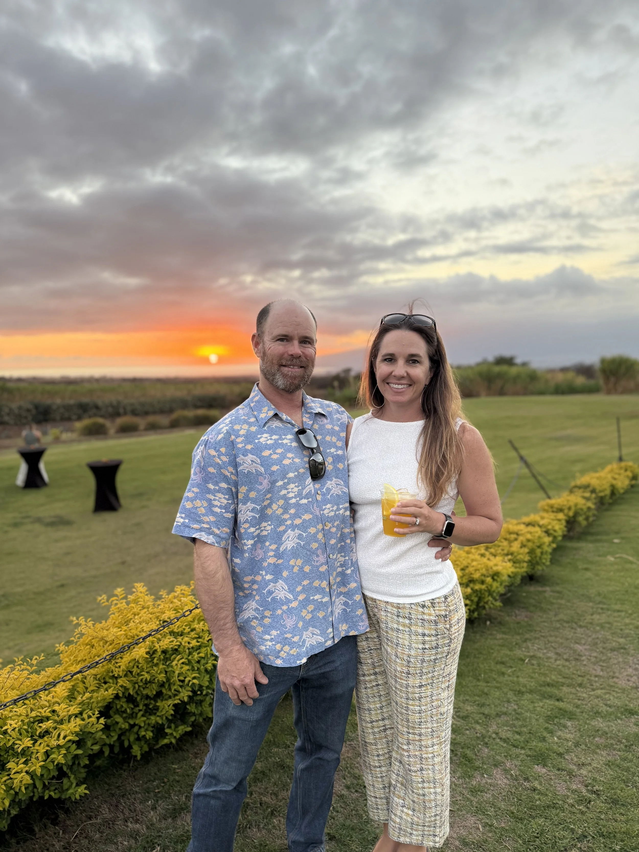 A man and woman smiling outdoors at sunset, standing next to a row of yellow bushes. The man has a beard and is wearing a blue Hawaiian shirt with sunglasses hanging from the collar. The woman has long brown hair, glasses on her head, and is holding a yellow drink.
