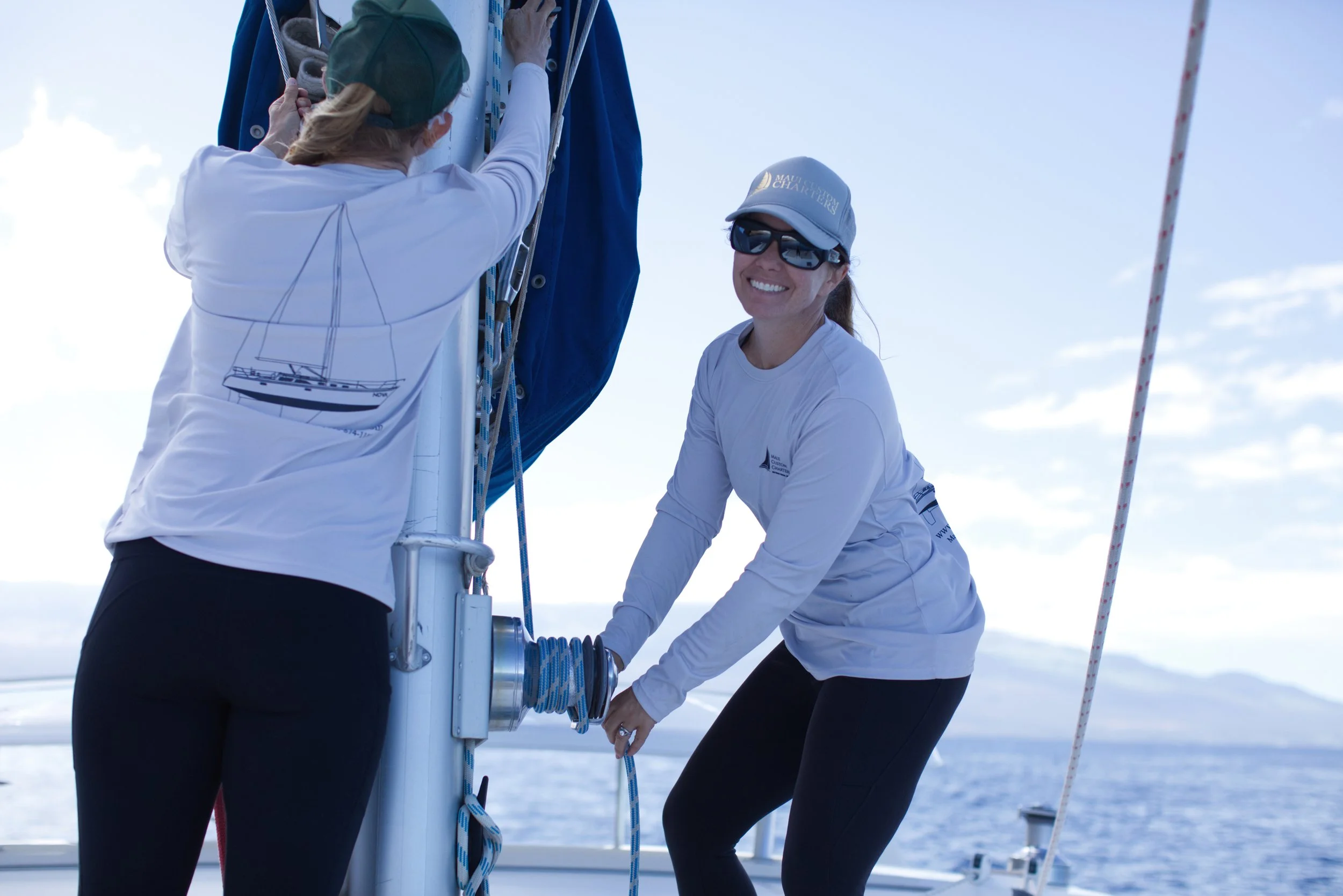 Two women are preparing a sailboat for sailing on a bright day. One woman is adjusting the rigging while the other is smiling and holding a rope. Both are dressed in sailing attire with hats and sunglasses, with the ocean and sky in the background.