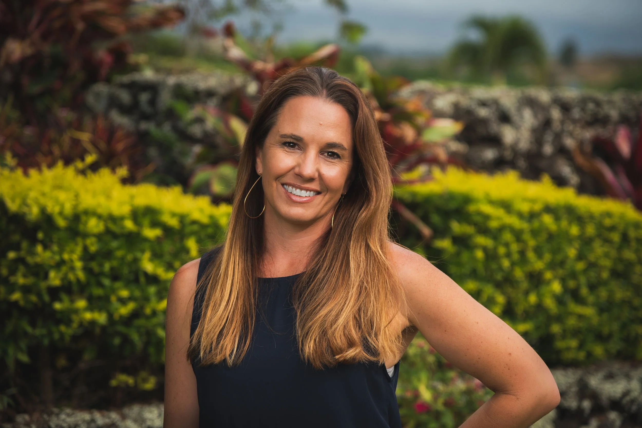 A woman with long brown hair smiling outdoors with colorful plants and a stone wall in the background.