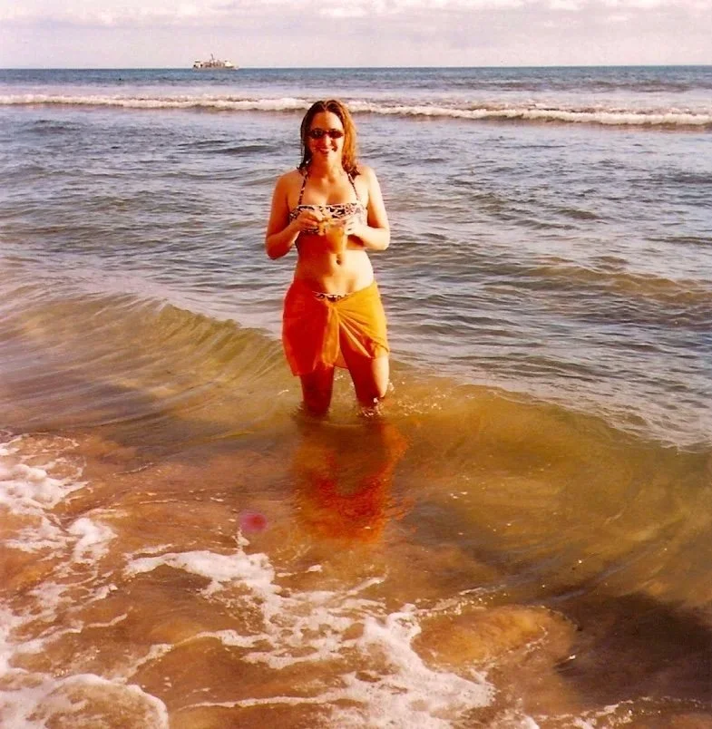 A woman standing in the ocean water at the beach, holding a drink, wearing sunglasses, a leopard print bikini top, and a yellow sarong. There is a ship in the distance.