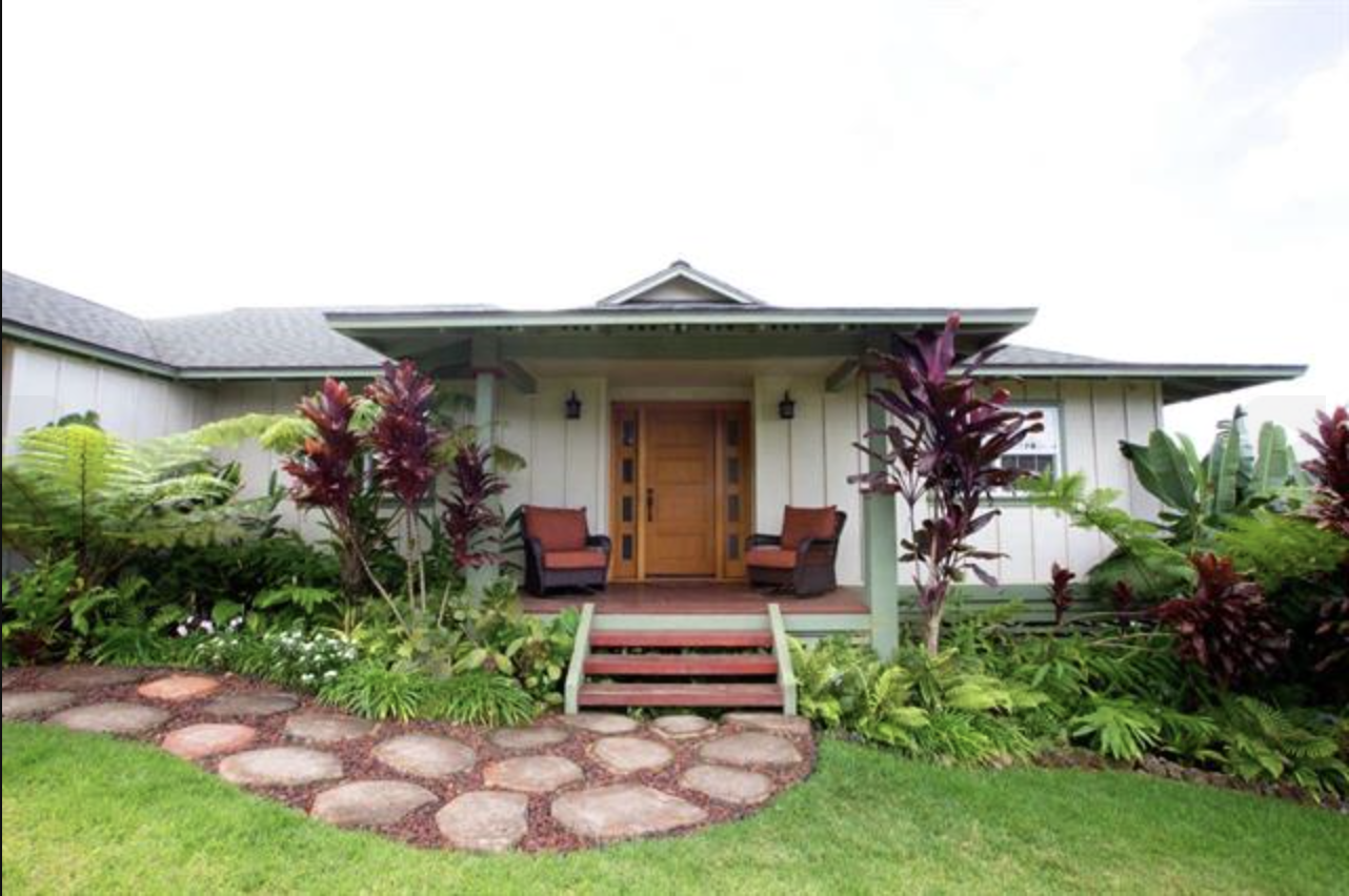Front view of a house with a wooden front door, two chairs on the porch, surrounded by lush green plants and red-leafed foliage, with a stone pathway leading to the stairs.