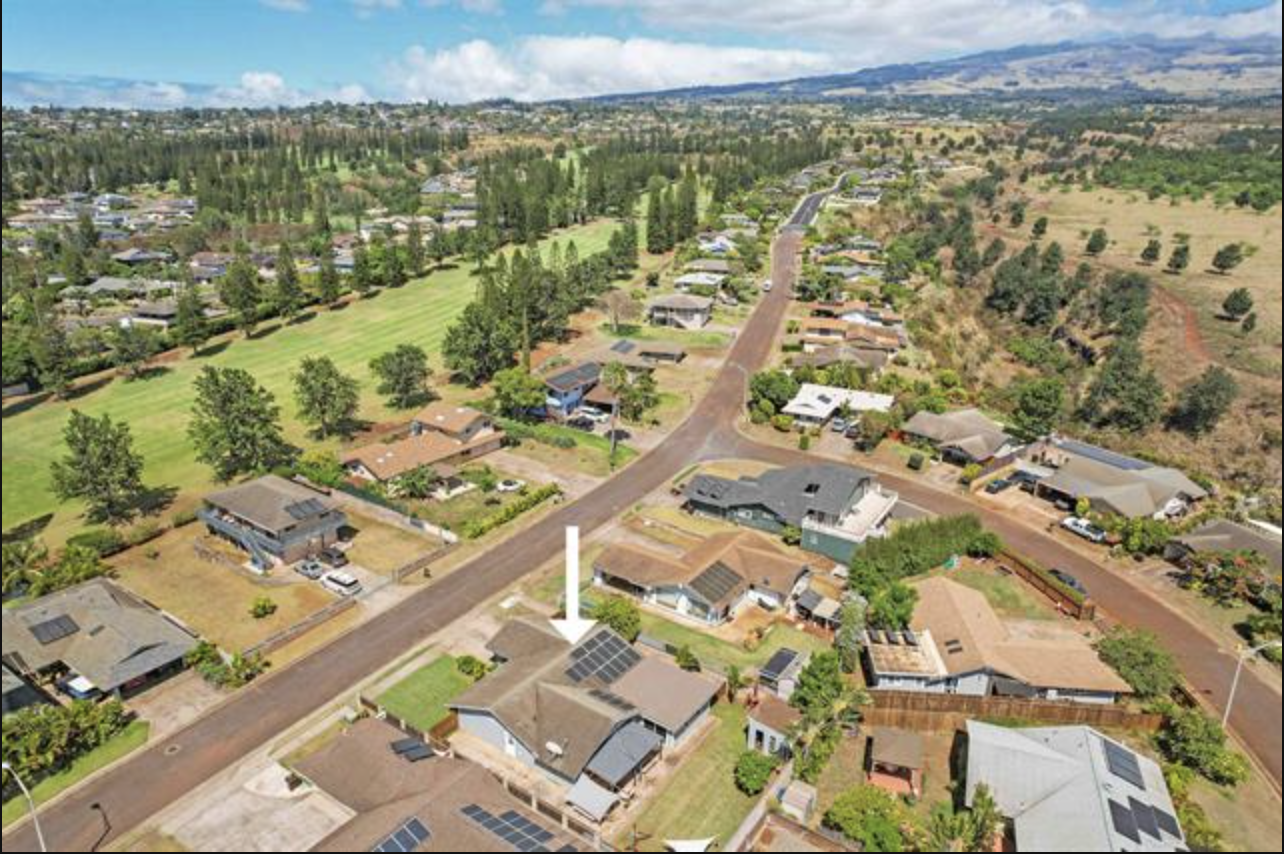 Aerial view of a suburban neighborhood with houses, trees, and a road with solar panels on the roof of one house, marked by an arrow.