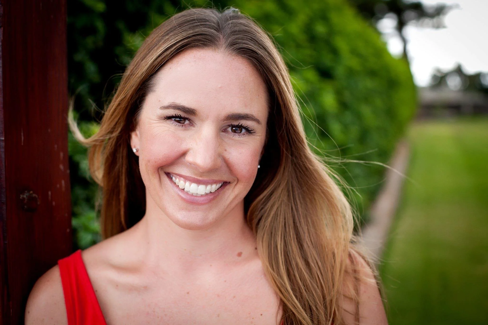 Close-up of a smiling woman with long brown hair, wearing a red tank top, standing outdoors with green foliage in the background.