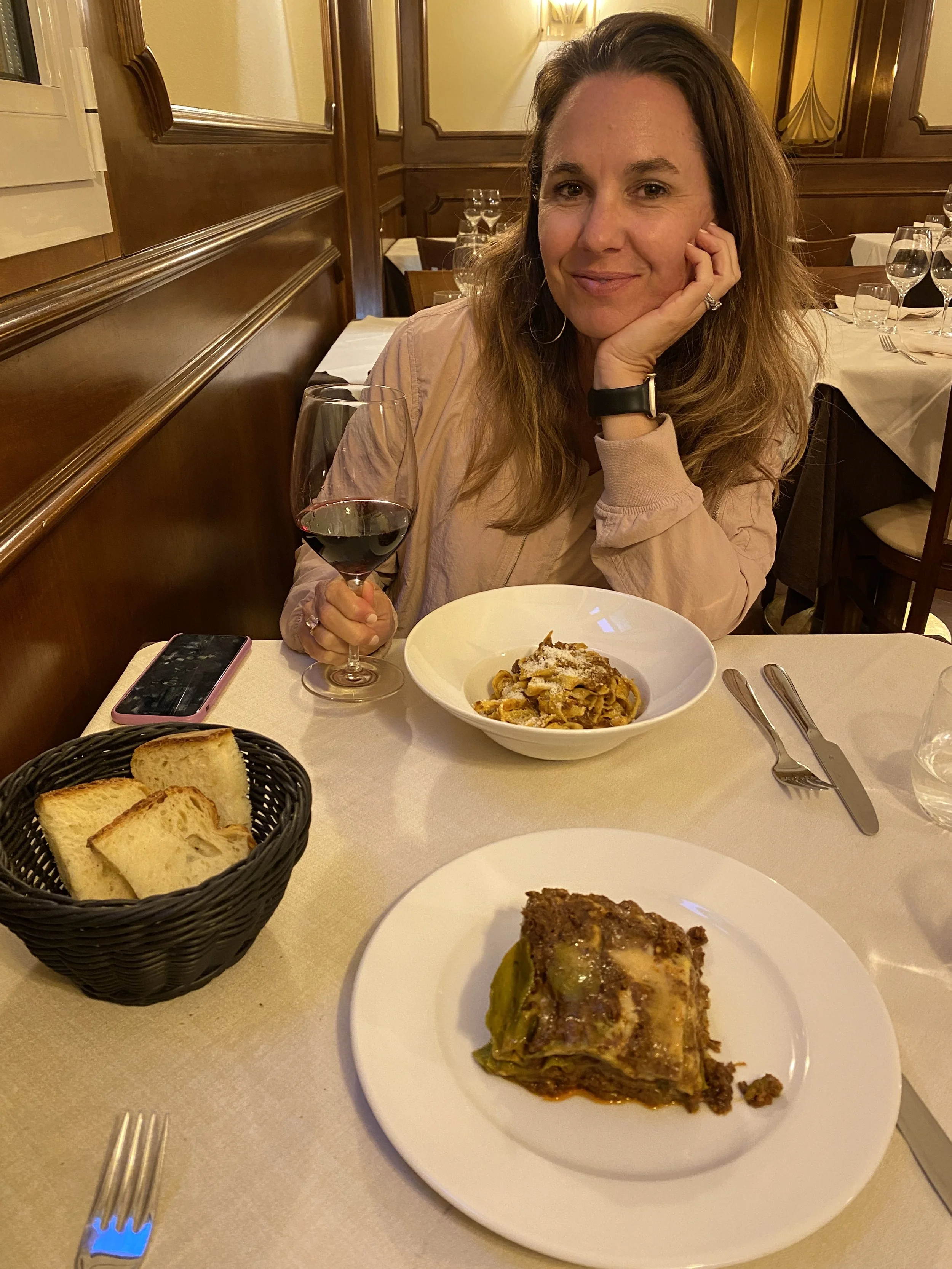 A woman with brown hair, wearing a beige jacket, smiling while sitting at a restaurant table. She has a glass of red wine in front of her and a plate of lasagna. The table has a basket of bread, utensils, and a smartphone. The restaurant has wood-paneled walls and neatly arranged tables with white tablecloths and glassware.