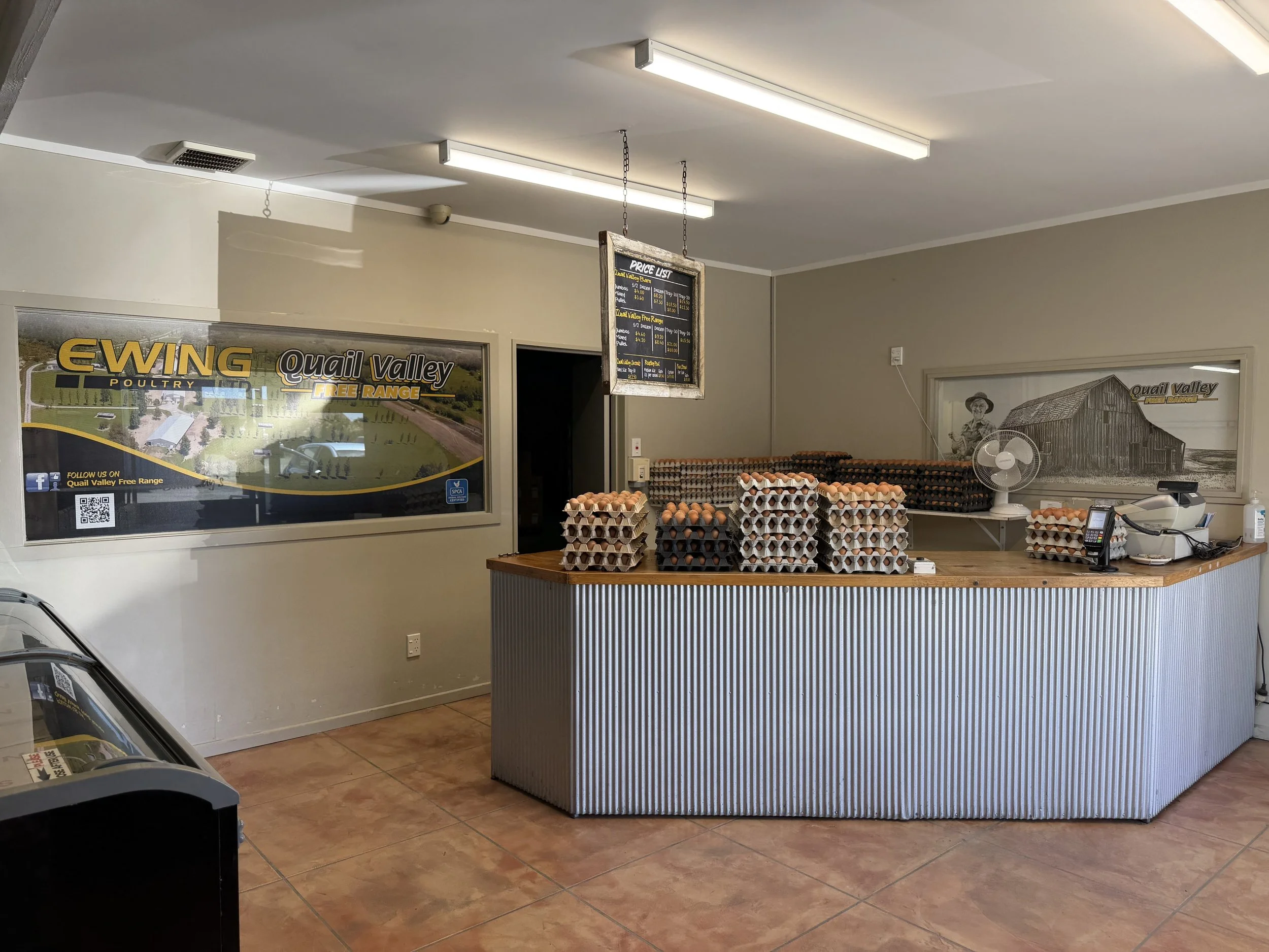 Interior of a poultry store displaying stacks of eggs on a counter, signage for Quail Valley Free Range Poultry, and a price list hanging from the ceiling. There is a fan, a cash register, and a framed black-and-white picture of a barn on the wall.