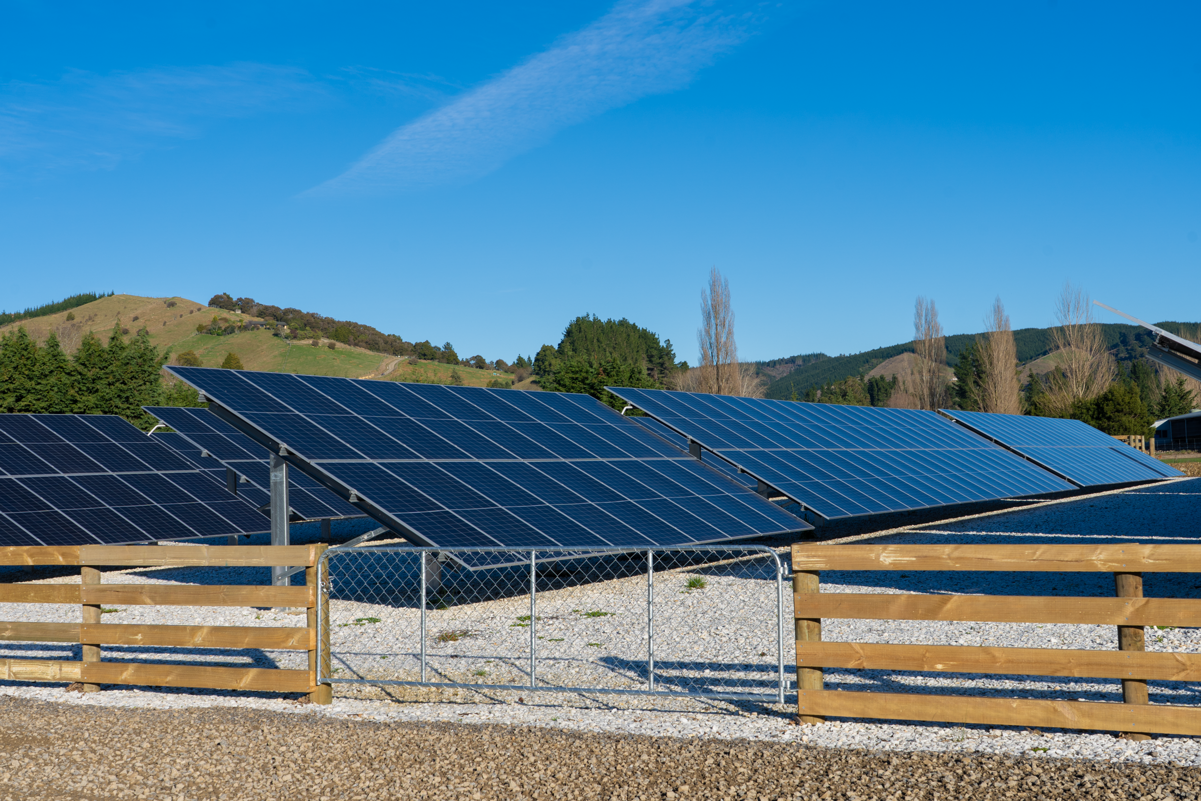 Solar panels installed on a gravel surface with a wooden fence in the foreground, green hilly landscape with trees and blue sky in the background.