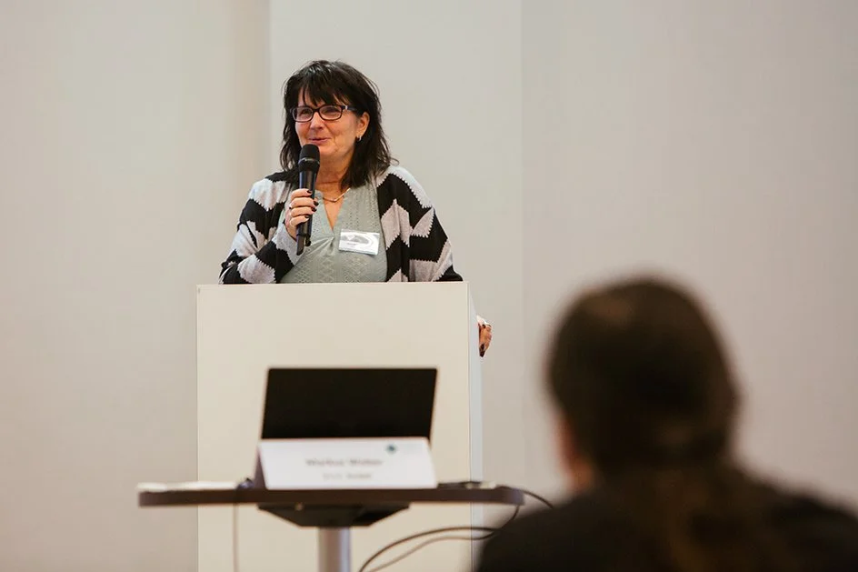 Eine Frau spricht an einer Konferenz oder Veranstaltung, steht hinter einem Rednerpult und hält ein Mikrofon, während sie einem Publikum präsentiert.