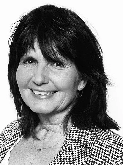 Schwarz-Weiß-Fotografie einer lächelnden Frau mit dunklem, glattem Haar und einem karierten Blazer.