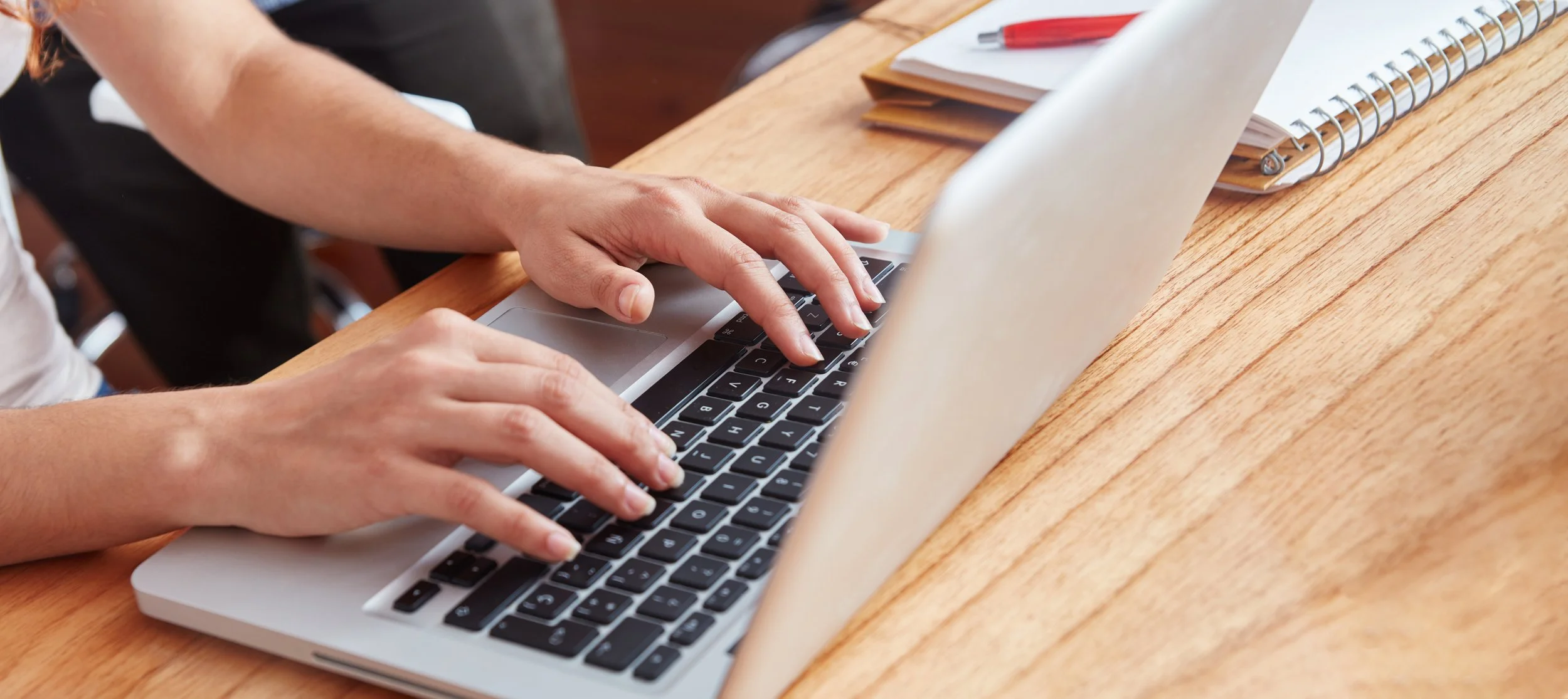 Person tippt auf einer silbernen Laptop-Tastatur, auf einem Holztisch mit Notizbüchern und einem roten Stift.