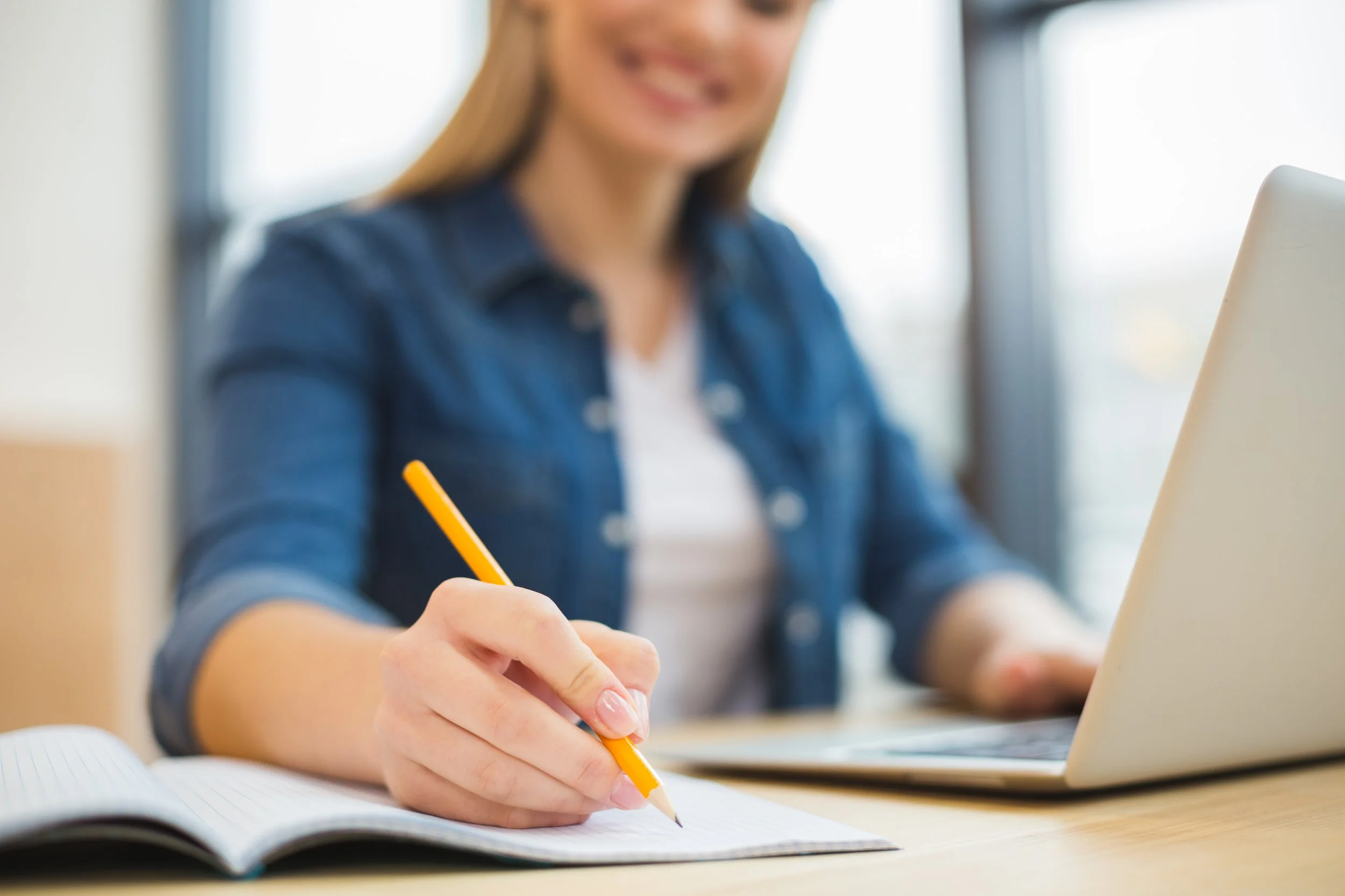 Eine junge Frau schreibt in einem Notizbuch mit einem Gelstift, während sie an einem Laptop arbeitet.