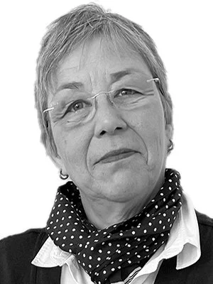 Porträt einer älteren Frau mit kurzen Haaren, Brille und einem schwarzen Pullover mit weißen Punkten, die einen weißen Hemdkragen trägt.